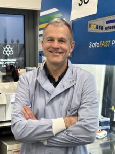 A man in a grey blue lab coat smiling towards the camera with his arms folded confidently.