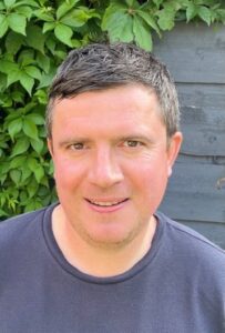 A portrait of man in a navy blue top smiling towards the camera, a green plant trails againt the wall behind him..